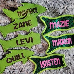 Puede incluir: Una colección de etiquetas con nombres personalizados en forma de pez. Las etiquetas son en tonos verdes, con nombres como "Maverick", "Zane", "Levi" y "Quill" en purpurina marrón, y "Mazie", "Madison" y "Kristen" en purpurina rosa.