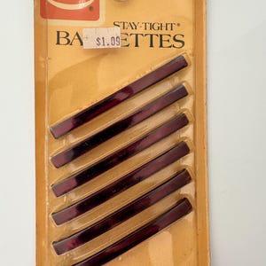 May include: A package of six Goody Stay-Tight barrettes. The barrettes are a deep burgundy color and are rectangular in shape. The packaging is a tan color with the Goody logo in the upper left corner. The package reads "6 BARRETTES MADE IN U.S.A."