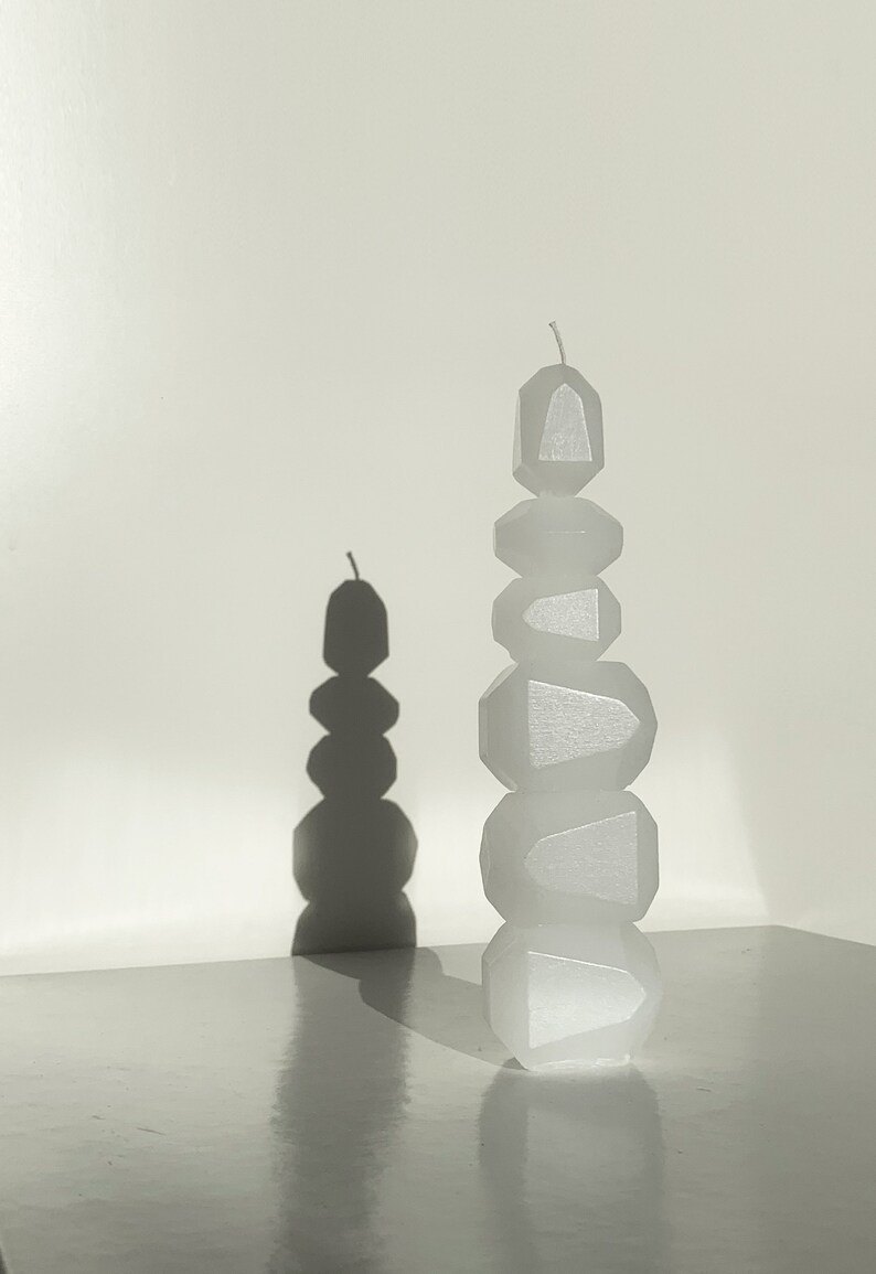 K&ouml;nnte beinhalten: Eine wei&szlig;e Kerze in Form eines Stapels geometrischer Formen. Die Kerze steht auf einer wei&szlig;en Oberfl&auml;che und wirft einen Schatten an die Wand dahinter.