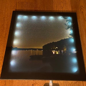Könnte beinhalten: Ein schwarzer gerahmter Leinwanddruck mit einer Nachtszene eines Sees mit einem Wald im Hintergrund. Die Leinwand ist mit weißen LED-Lichtern um den Umfang beleuchtet.