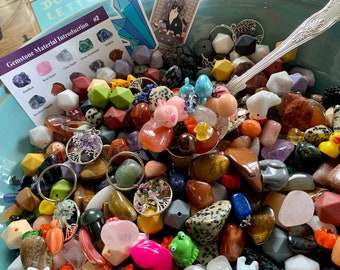 May include: A bowl filled with a variety of colorful gemstones, beads, and small figurines. The bowl is made of light blue ceramic and has a silver spoon resting on top of the gemstones. The gemstones include amethyst, rose quartz, clear quartz, green aventurine, sodalite, and carnelian. The beads are made of various materials, including wood, glass, and plastic. The figurines include a pig, a frog, a duck, and a unicorn. The image also includes a card with the text "Gemstone Material Introduction #2".