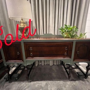 May include: A vintage wooden buffet with a dark wood finish and green paint. The buffet has two doors on each side and three drawers in the center. The buffet is sitting on a light gray floor.