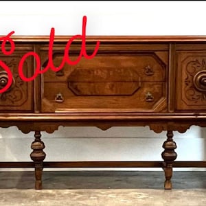 May include: A vintage wooden buffet with carved details and turned legs. The buffet has two doors on the right and two drawers on the left. The top is a rich brown color and the legs are a darker brown. The buffet is in good condition and would be a great addition to any home.