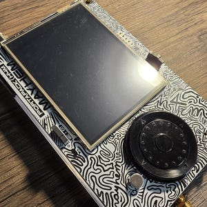 May include: A white and black patterned electronic device with a black and white screen, a black knob, and a silver button. The device has the text "PORTAPACK.COM" and "MATHEN" printed on it.