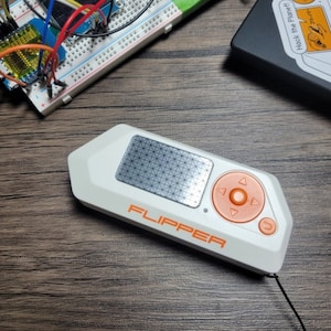 May include: A white and orange Flipper device with a black strap. The device has a small screen with a grid pattern and a circular button with directional arrows. The text "FLIPPER" is printed in orange on the device.