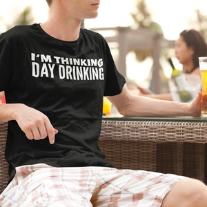 May include: A person wearing a black t-shirt with the text "I'M THINKING DAY DRINKING" in white letters. The person is sitting at a table with a glass of beer in their hand.