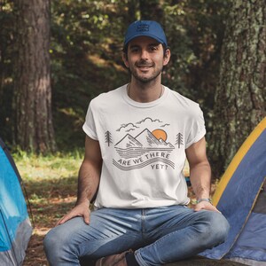 Op de afbeelding: Een wit T-shirt met een afbeelding van bergen, een zon en bomen. De tekst "Are we there yet?" staat op het T-shirt gedrukt. Een persoon die het T-shirt draagt, zit op een boomstam met een hond.