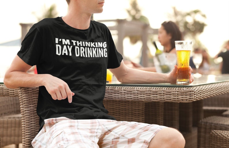 May include: A person wearing a black t-shirt with the text "I'M THINKING DAY DRINKING" in white letters. The person is sitting at a table with a glass of beer in their hand.