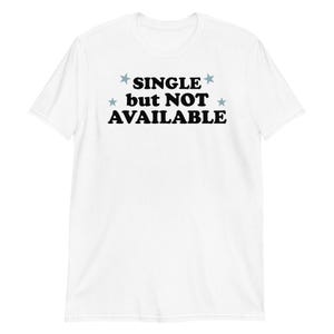 May include: A white short-sleeve t-shirt featuring the black text "SINGLE but NOT AVAILABLE" printed on the chest. The text is flanked by four small, light blue stars arranged in a symmetrical pattern above and below the words.