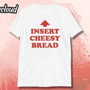May include: White t-shirt with the text "INSERT CHEESY BREAD" in red, with a red arrow pointing upwards. The shirt has a crew neck and short sleeves. The brand name "Teecloud" is in the upper left corner.