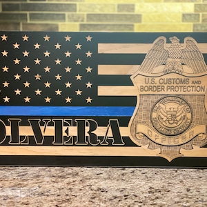May include: Wooden American flag with a blue line and the name "OLVERA" engraved. Features a U.S. Customs and Border Protection badge. The flag has black and natural wood stripes with stars in the upper left corner.