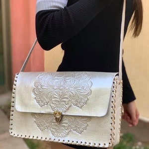 May include: A white leather shoulder bag with a gold clasp and intricate floral embossed design. The bag has a long strap and a stitched border.