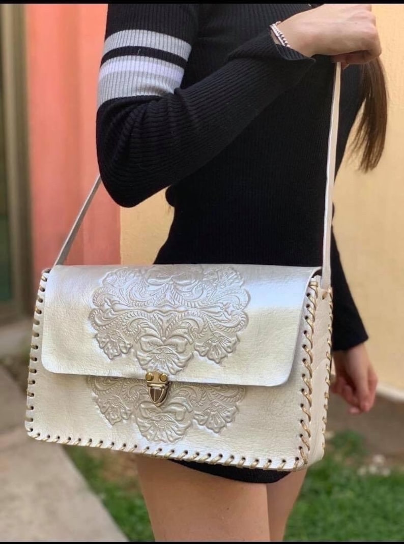May include: A white leather shoulder bag with a gold clasp and intricate floral embossed design. The bag has a long strap and a stitched border.