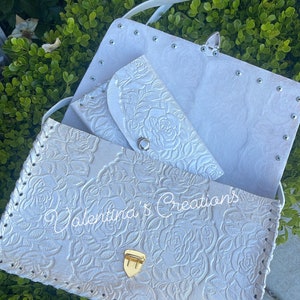 May include: White leather purse with a floral embossed design and a gold clasp. The purse has a matching smaller wallet inside and a white strap.