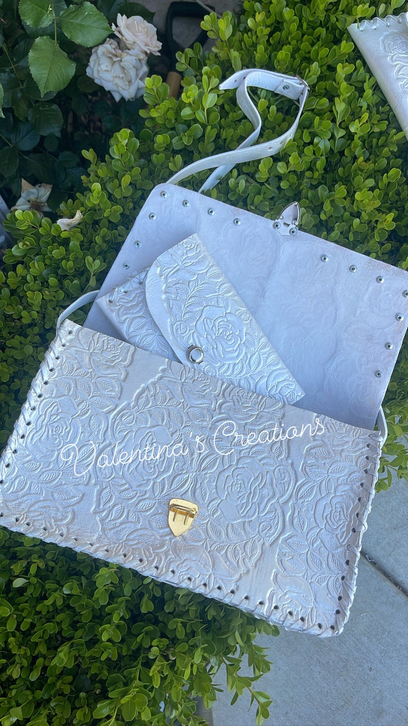 May include: White leather purse with a floral embossed design and a gold clasp. The purse has a matching smaller wallet inside and a white strap.