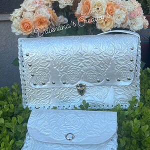 May include: White leather clutch purse with a rose embossed design and a gold clasp. The purse has a matching smaller wallet with a snap closure.