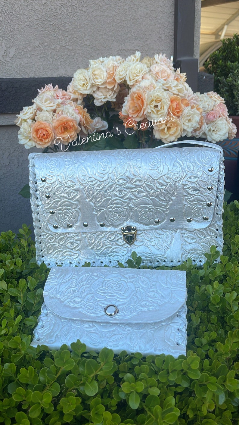 May include: White leather clutch purse with a rose embossed design and a gold clasp. The purse has a matching smaller wallet with a snap closure.