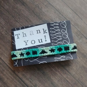 May include: A black and green friendship bracelet with a white background. The bracelet is on a card that says "Thank You!"