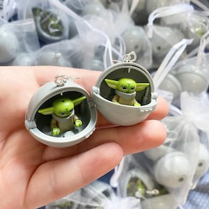 May include: A pair of silver earrings with a small, green, and gray figurine of a character from the Star Wars franchise inside a sphere. The earrings are displayed on a hand against a background of white mesh bags.