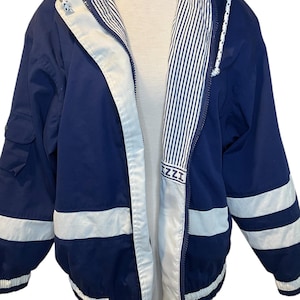 Puede incluir: Chaqueta azul marino con detalles blancos y forro a rayas. La chaqueta tiene capucha, cierre de cremallera y la marca "IZZI". Rayas blancas en las mangas y el dobladillo.