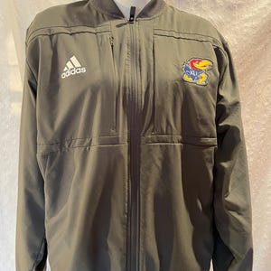 May include: A dark gray Adidas jacket with a zipper closure and a small Adidas logo on the left chest. The jacket has a KU logo patch on the right chest.