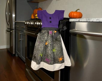 Peut inclure: Un torchon de cuisine sur le thème d'Halloween en forme de robe. Le haut est violet avec des boutons noirs, et la jupe présente un motif de toile d'araignée avec des araignées orange, vertes et violettes. Il y a un nœud noir attaché.