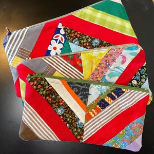 May include: Three colorful, patchwork zipper pouches in varying sizes. Each pouch features a unique combination of fabrics, including floral, striped, and solid patterns in red, green, blue, and brown. The pouches have metal zippers.