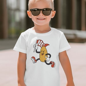 Puede incluir: Camiseta blanca con un dibujo animado de una mazorca de maíz sonriente, sosteniendo una bandera con "NE". El maíz está corriendo y lleva zapatos rojos. La camiseta la lleva un niño.