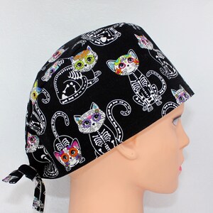 Puede incluir:  gorro de quir&oacute;fano negro con un estampado de gato del D&iacute;a de los Muertos colorido. Los gatos tienen esqueletos blancos y flores coloridas en sus caras.
