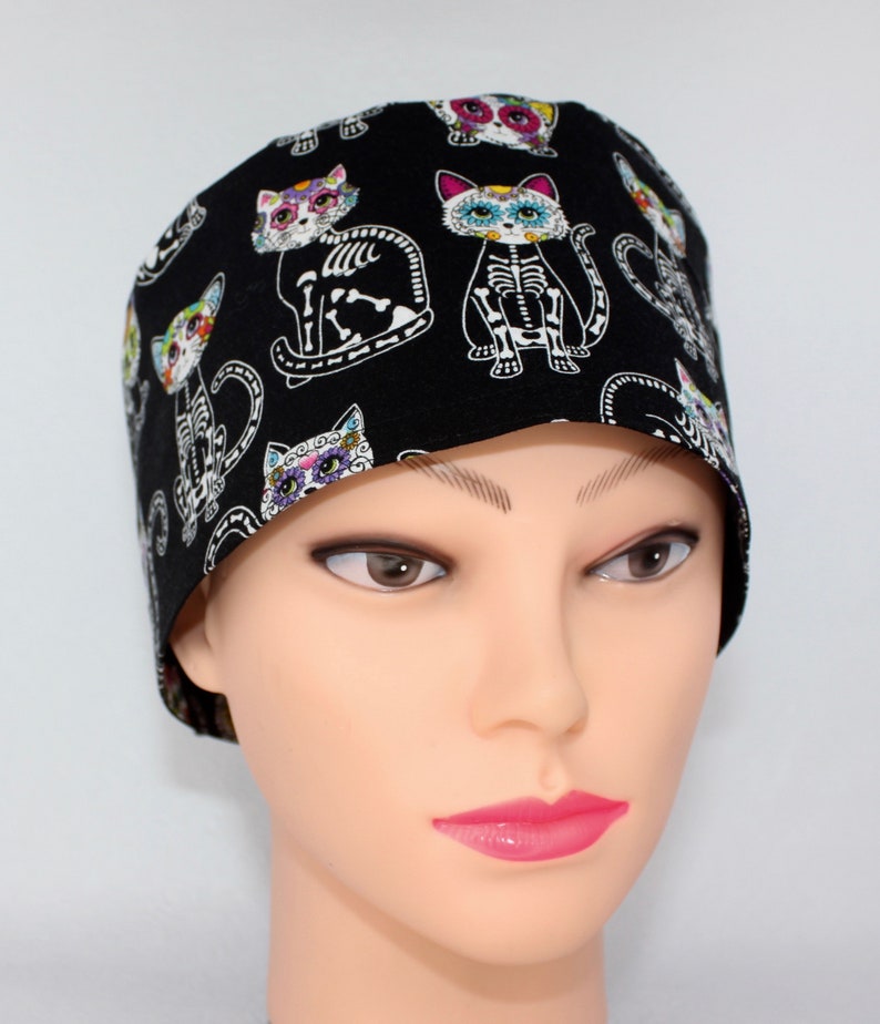 Puede incluir:  gorro de quir&oacute;fano negro con un patr&oacute;n de gato colorido del D&iacute;a de los Muertos. El gato tiene un dise&ntilde;o de esqueleto y detalles florales.