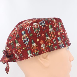 May include: A scrub cap featuring a deep red background with a repeating pattern of colourful nutcracker figures and small gold stars. The cap has a tie-back closure at the nape of the neck, providing a secure fit with a rounded crown design.