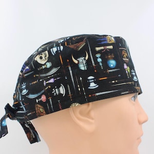 May include: Black surgical scrub cap with a repeating pattern of fantasy weapons, including swords, axes, and scrolls. The cap is tied at the back and is designed to cover the hair during medical procedures or other activities.