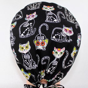 Puede incluir:  gorro de quir&oacute;fano negro con un patr&oacute;n repetido de gatos de calavera de az&uacute;car blancos con flores de colores. El gorro tiene lazos negros.