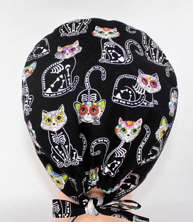 Puede incluir:  gorro de quir&oacute;fano negro con un patr&oacute;n repetido de gatos de calavera de az&uacute;car blancos con flores de colores. El gorro tiene lazos negros.