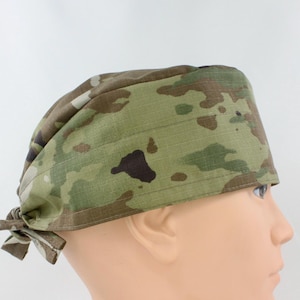 May include: A scrub cap featuring a multi-colored camouflage pattern in shades of olive green, brown, tan, and black. The fabric has a visible grid-like texture and includes ties at the back to secure the fit around the head for a snug appearance.