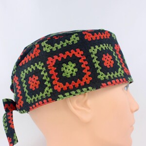 May include: A scrub cap featuring a black background with a repeating pattern of red and green square motifs. The cap has a fitted crown and a tie-back closure at the nape of the neck, constructed from fabric with a visible stitched texture.