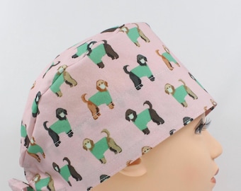 May include: A pink surgical scrub cap featuring a repeating pattern of dogs wearing green sweaters and face masks. The dogs are in various colors, including brown, black, and tan. The cap is tied at the back with a matching pink fabric.