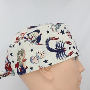 May include: A white scrub hat with a colorful tattoo-inspired print featuring a skull, a scorpion, a mermaid, dice, and stars.