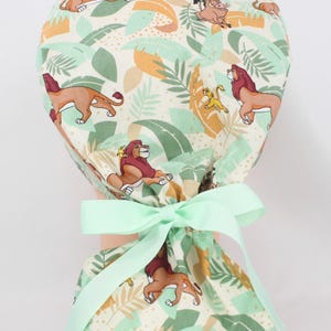May include: A scrub cap featuring a repeating pattern of lions, leaves, and other characters in shades of brown, yellow, and green on a cream background. The cap is tied with a light green ribbon.