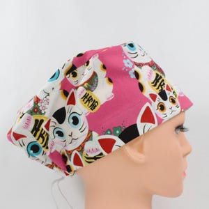 May include: A pink and white patterned surgical scrub cap featuring a repeating design of Japanese Maneki Neko lucky cats. The cats have black and red accents, with gold coin details. The cap is tied in the back.