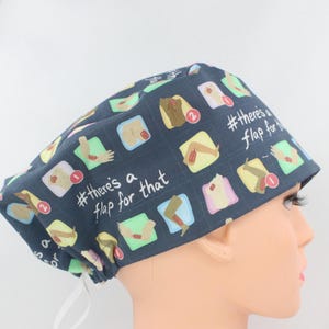 May include: A dark blue scrub cap featuring a repeating grid pattern of various human body parts with surgical flaps and red notification icons. The text #there's a flap for that is printed in white script across the fabric surface.
