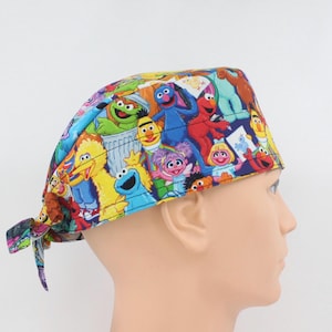 May include: A colorful surgical scrub cap featuring a vibrant print of Sesame Street characters. The cap is tied at the back and is made of a lightweight fabric. The design includes characters like Big Bird, Cookie Monster, and Elmo.