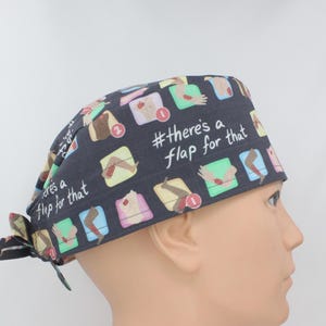 May include: A dark gray surgical scrub cap with a repeating pattern of medical illustrations and the text "#there's a flap for that" in white. The cap is tied at the back and is worn by a mannequin.