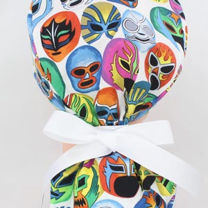 May include: A white scrub cap featuring a vibrant pattern of colorful luchador masks. The masks are in various colors, including red, blue, yellow, and green. A white ribbon is tied in a bow at the back.