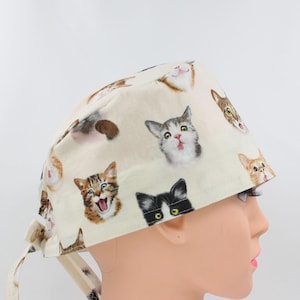 Peut inclure: Une toque chirurgicale crème avec un motif répété de visages de chats de différentes couleurs et expressions. La toque a des liens à l'arrière pour un ajustement réglable.