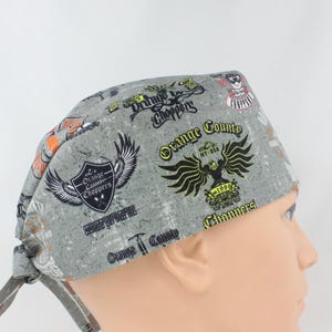 May include: A gray scrub cap featuring a repeating pattern of Orange County Choppers logos, eagle emblems, and text including Orange County Choppers, NY-USA, and est. 1999. The fabric has a distressed, textured appearance with black, yellow, and white graphics.