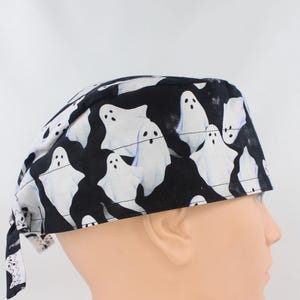 May include: A black scrub cap featuring a repeating pattern of white ghosts with black eyes and open mouths. The fabric head covering has a rounded crown and ties at the back, providing a secure fit for medical or professional use.