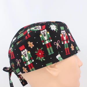 May include: A black scrub cap featuring a festive holiday print with nutcracker figures, gingerbread men, candy canes, poinsettias, and white snowflakes. The cap has a tie-back closure and is constructed from fabric with a repeating pattern.