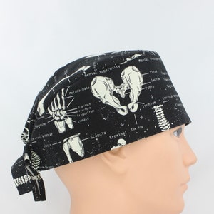 May include: Black scrub hat with a white anatomical illustration of the human skeleton, including the hip, spine, and hand bones. The hat has a tie on the back.