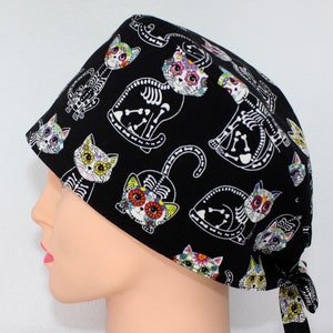 Puede incluir:  gorro de quir&oacute;fano negro con un estampado de gato colorido del D&iacute;a de Muertos. Los gatos tienen calaveras de az&uacute;car y dise&ntilde;os florales.